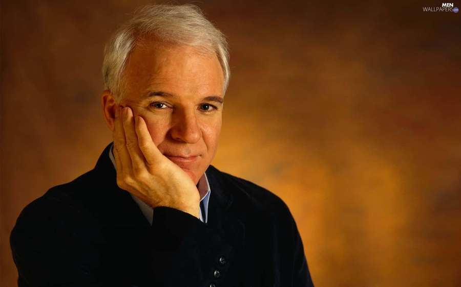 A man with white hair in a dark blazer rests his chin on his hand with a gentle smile. — Steve martin Wallpaper