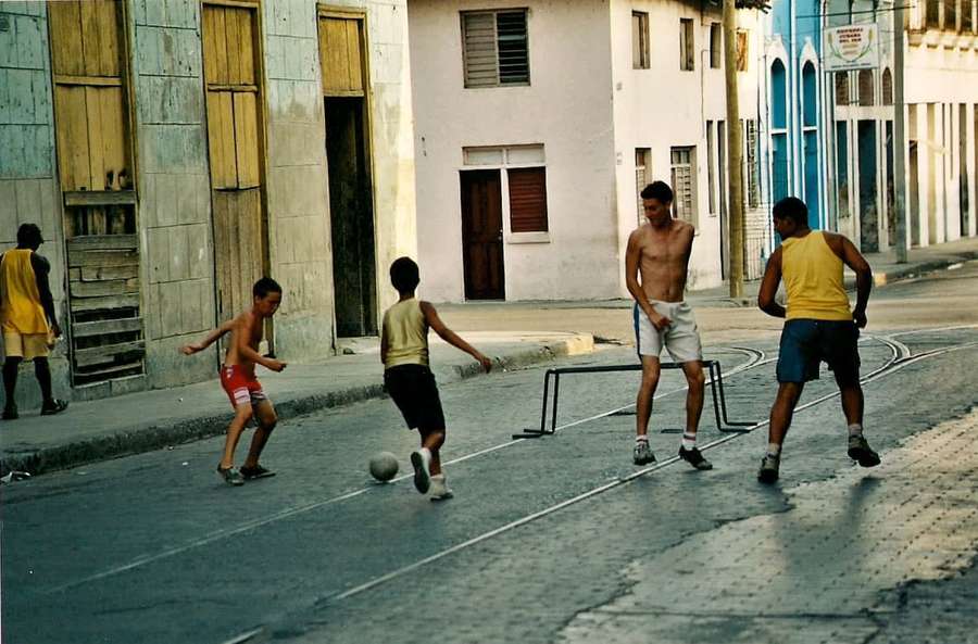 Children and adults playing soccer on a sunlit city street with old buildings. — Street Soccer Wallpaper
