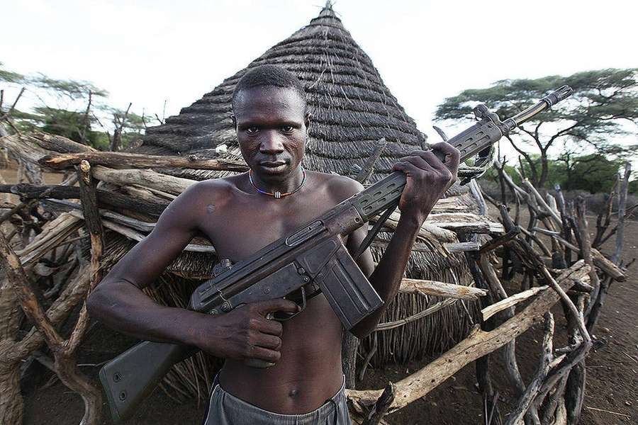 Um homem segurando um rifle em frente a uma cabana de palha em um ambiente rural africano. — Sudão do Sul Papéis de Parede
