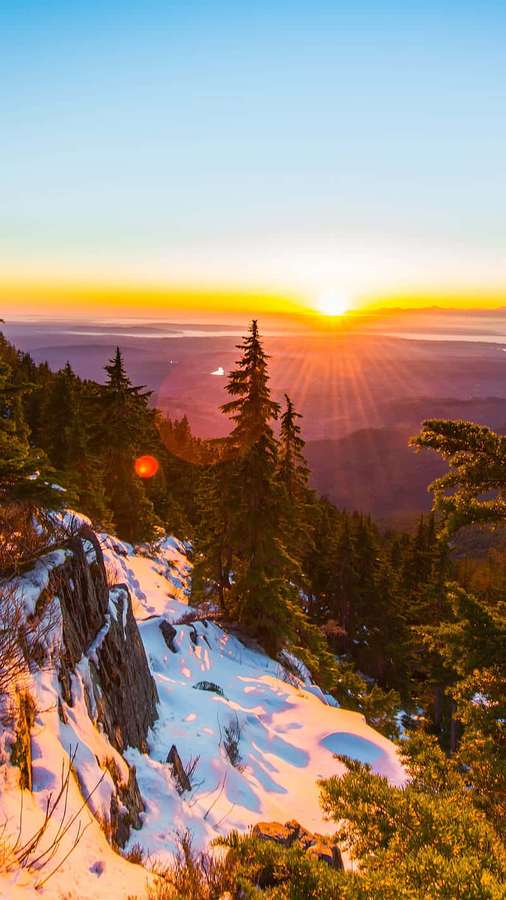 Snowy mountain ridge with evergreens at sunset casting golden light over a valley. — Sunrise iPhone Wallpaper