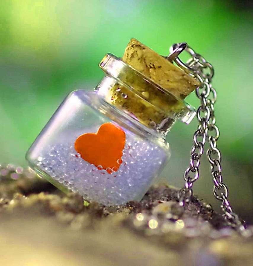 Eine kleine Glasflasche mit Korken und Kette enthält ein leuchtend orangefarbenes Herz und blaue Perlen auf sandigem Boden. — Süße Liebe Wallpaper