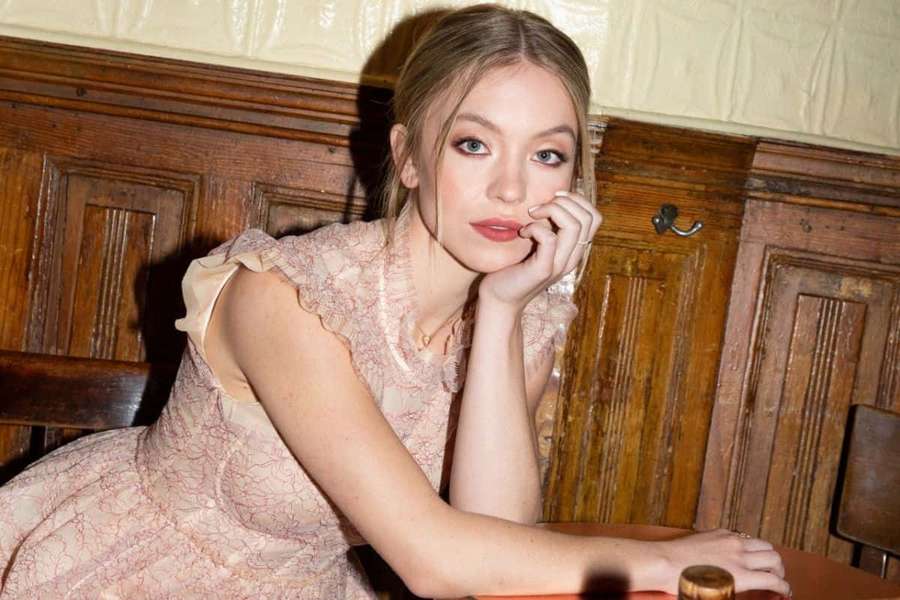 Young woman in pink lace dress resting chin on hand at wooden table indoors — Sydney Sweeney Wallpaper
