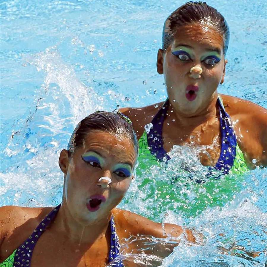 Two synchronized swimmers with colorful makeup and nose clips splashing in a pool. — Synchronized Swimming Background