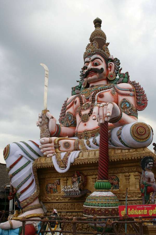 Large seated deity statue with sword and staff in ornate attire under cloudy sky. — Tamil God Background