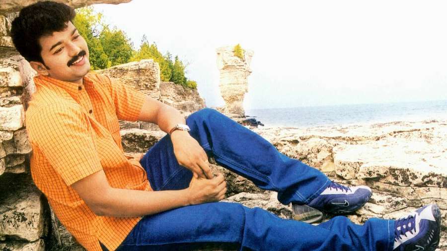 Man in orange shirt and blue jeans smiling on rocky seaside cliff. — Thalapathy HD Wallpaper