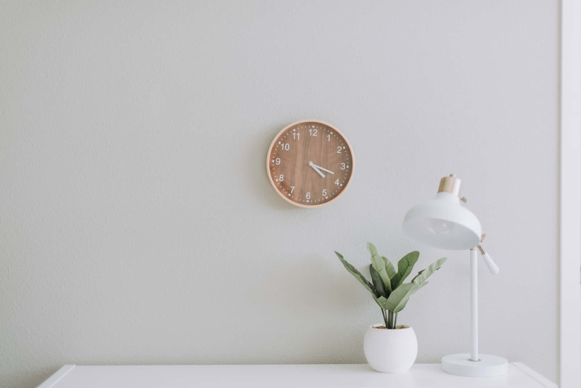 Vitt skrivbord med växt, lampa och träväggklocka i minimalistiskt rum. — Time Clock bakgrund Bakgrundsbilder