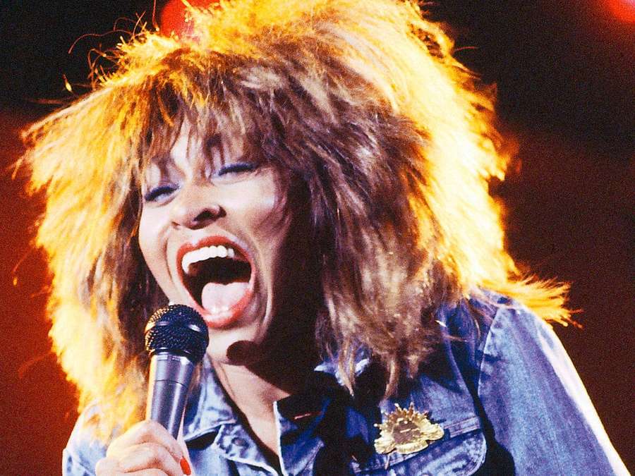 Cantante con capelli selvaggi che canta in un microfono sul palco. — Tina Turner Sfondi