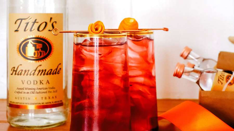 Zwei rote Cocktails mit Orangengarnitur und Eis neben einer Wodkaflasche auf Holz. — Titos Wodka Wallpaper