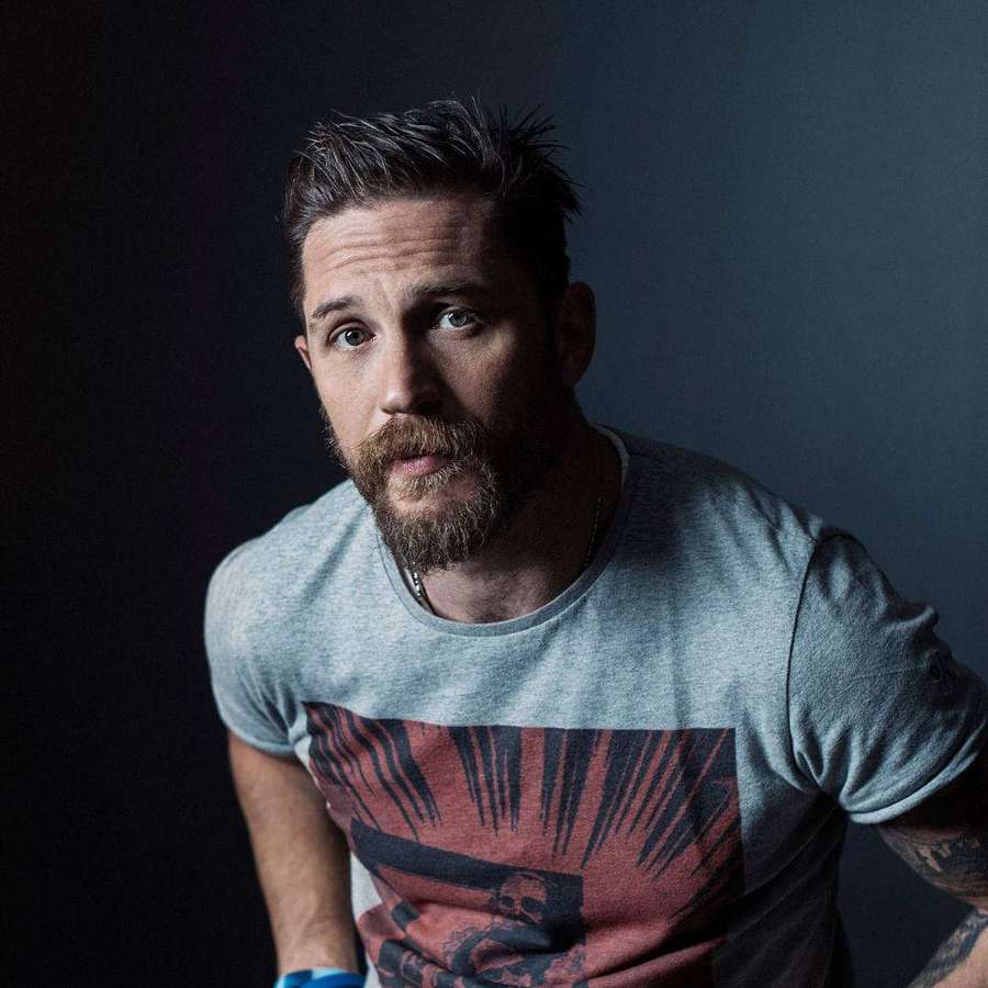A bearded man in a graphic t-shirt stares intensely at the camera in a dark studio setting. — Tom Hardy Background