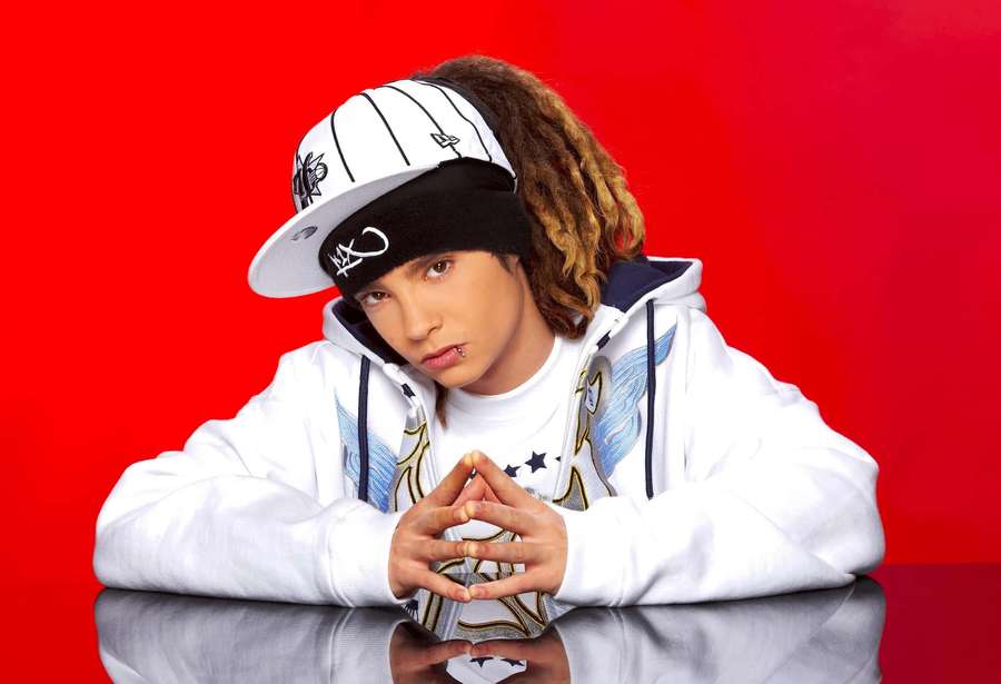 Person in white hoodie and cap leaning on reflective surface against red background. — Tom Kaulitz Wallpaper