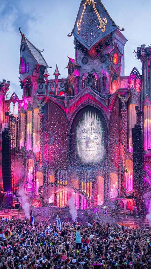 Crowd at a nighttime festival under a glowing castle stage with a projected face and confetti. — Tomorrowland Background