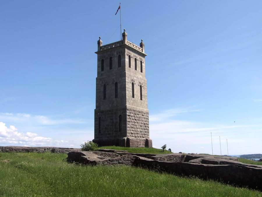 Stone tower on grassy hill with flag flying under blue sky — Tønsberg Wallpaper