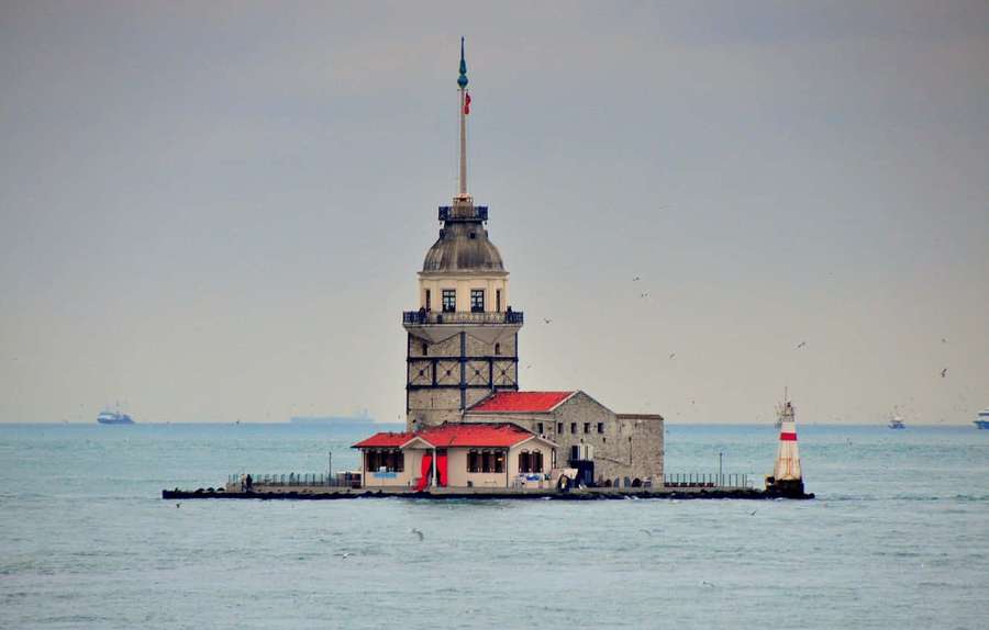 Torre de pedra com telhado vermelho e bandeira em pequena ilha no mar sob céu cinza — Torre da Donzela Papéis de Parede