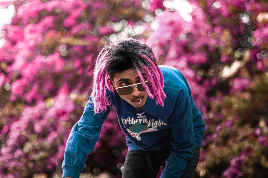 Jovem com dreadlocks rosa e óculos de sol posando ao ar livre entre flores rosa. — Tranças Papéis de Parede