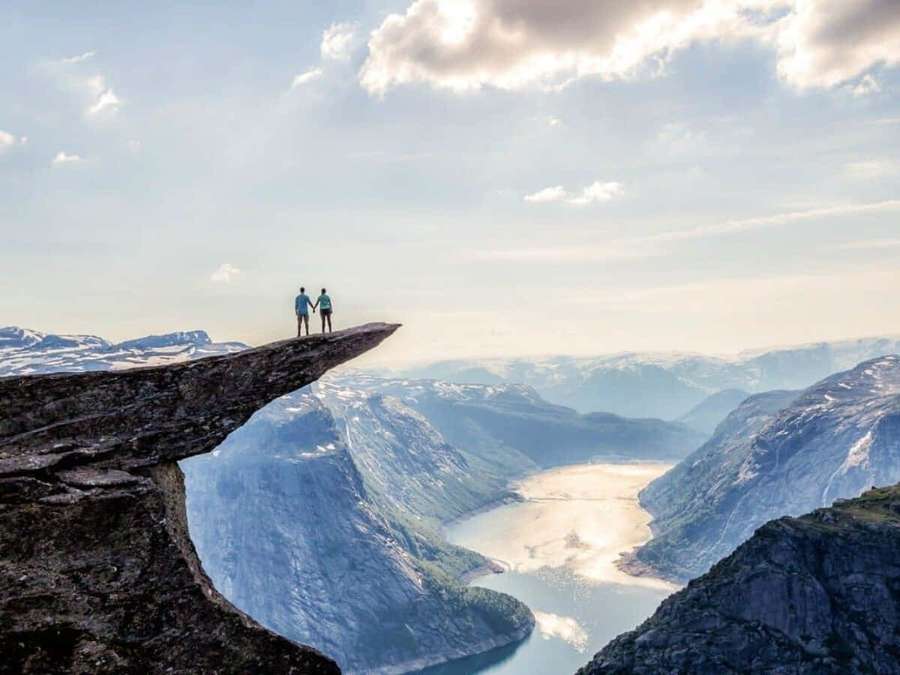 To personer holder hænder på en klippekant med udsigt over en fjord og bjerge ved solnedgang. — Trolltunga Wallpapers