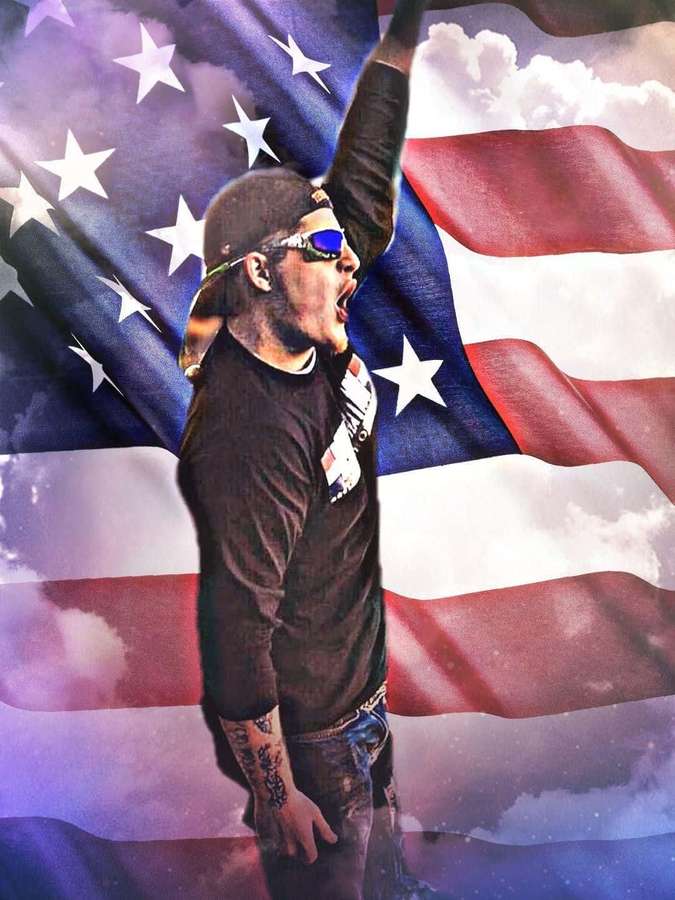 Hombre con tatuajes y gafas de sol gritando bajo una bandera estadounidense ondeante. — Upchurch Fondos de pantalla