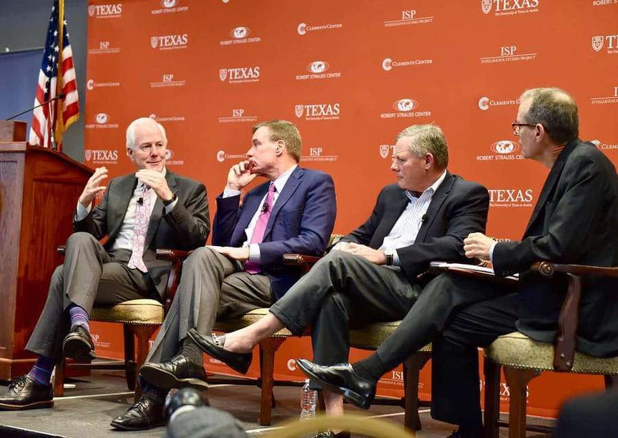 Four men in suits seated on stage at a university panel discussion. — USA Flag Wallpaper