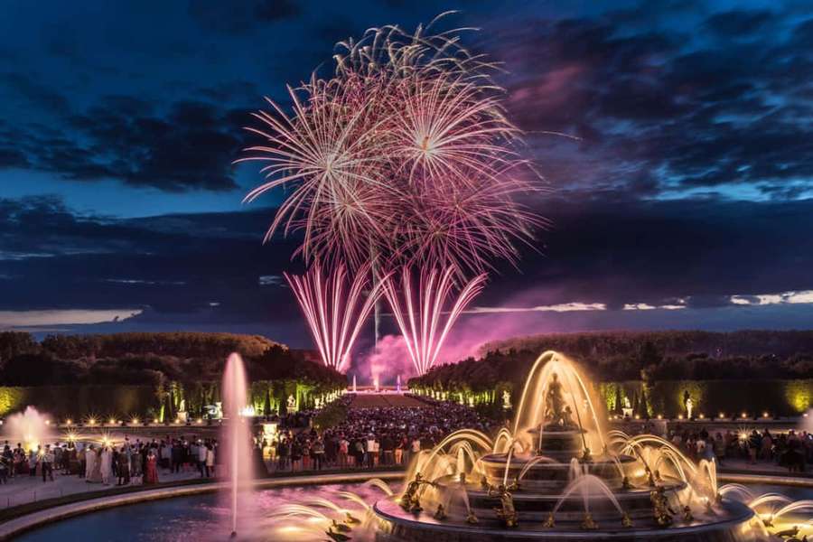 Fireworks burst over a lit fountain as people watch from a palace garden at night. — Versailles Wallpaper