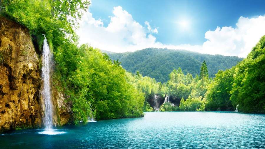 Uma cachoeira flui para um lago turquesa cercado por densa floresta verde sob um céu ensolarado. — Vista do rio Papéis de Parede