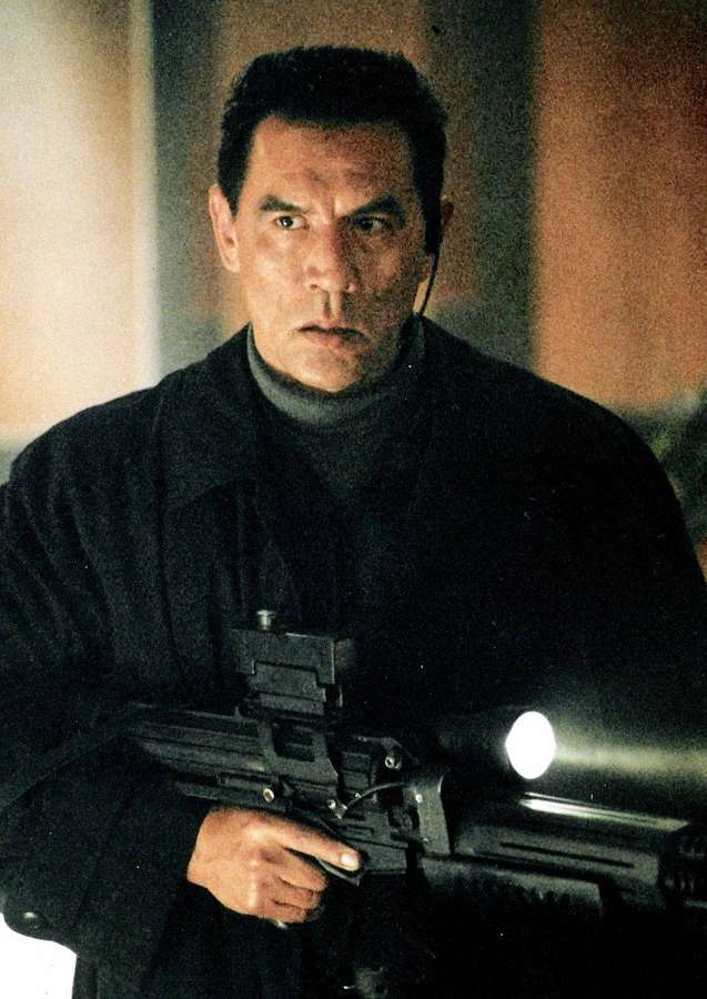 Man in dark coat holding rifle with flashlight indoors. — Wes Studi Background