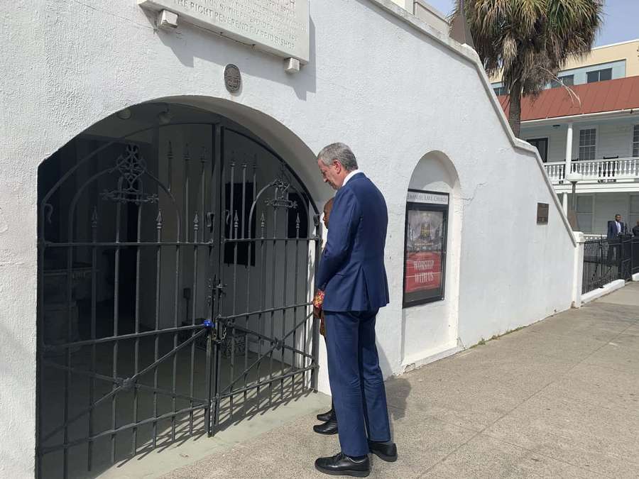 Homem de terno azul em pé no portão preto ornamentado de edifício histórico branco. — White Wall Papéis de Parede