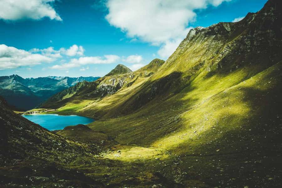 Sunlit mountain lake surrounded by green hills and distant peaks under a blue sky. — Winterthur Wallpaper