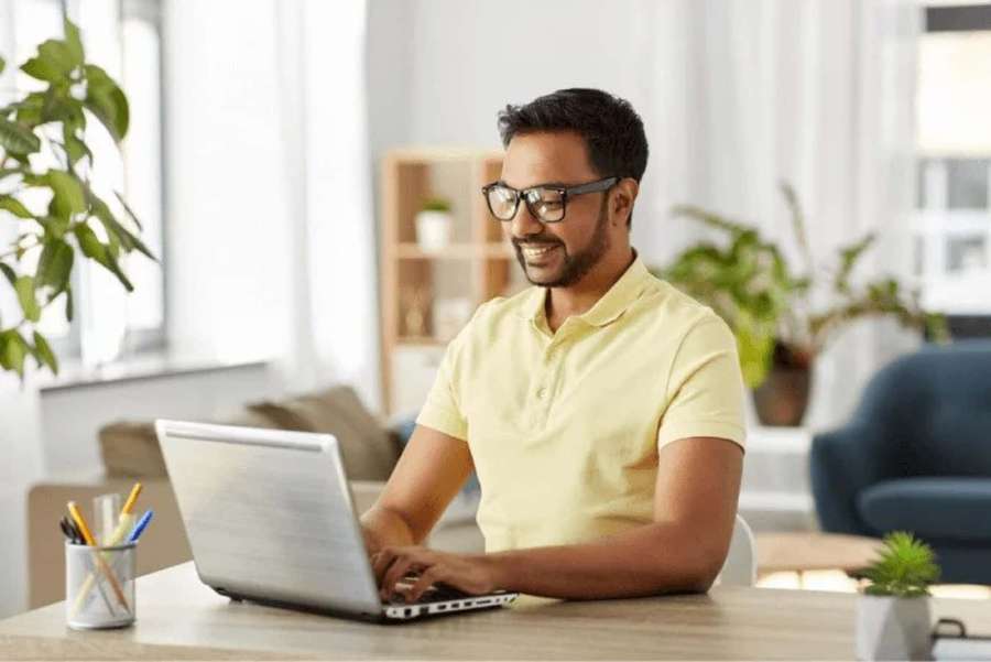Man wearing glasses smiles while typing on laptop at home desk with plants. — Work Picture