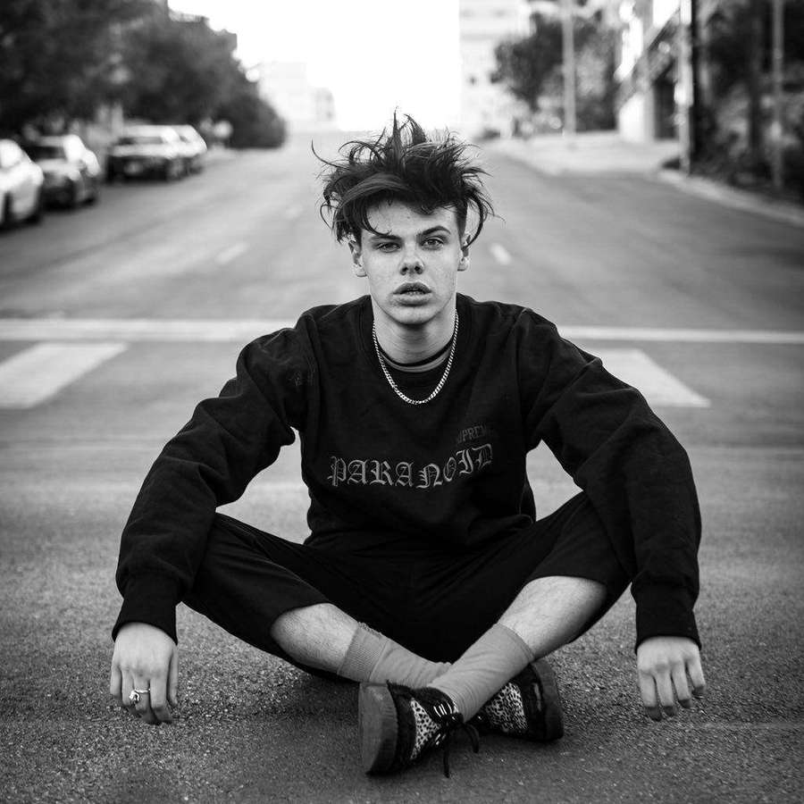 Young man sitting cross-legged on asphalt street wearing black sweatshirt and patterned sneakers. — Yungblud Wallpaper