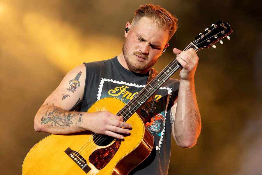 Male musician playing acoustic guitar on stage with tattoos under warm lights. — Zach Bryan Wallpaper