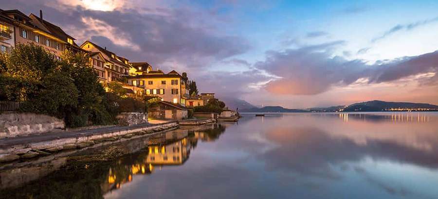Lakeside houses lit at dusk with reflections on calm water under twilight sky — Zug Wallpaper