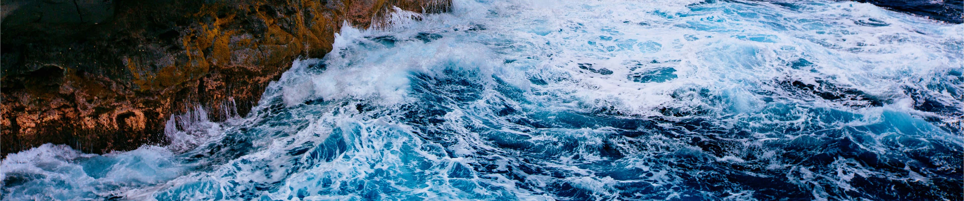 A Blue Ocean With Waves Crashing Over Rocks Wallpaper