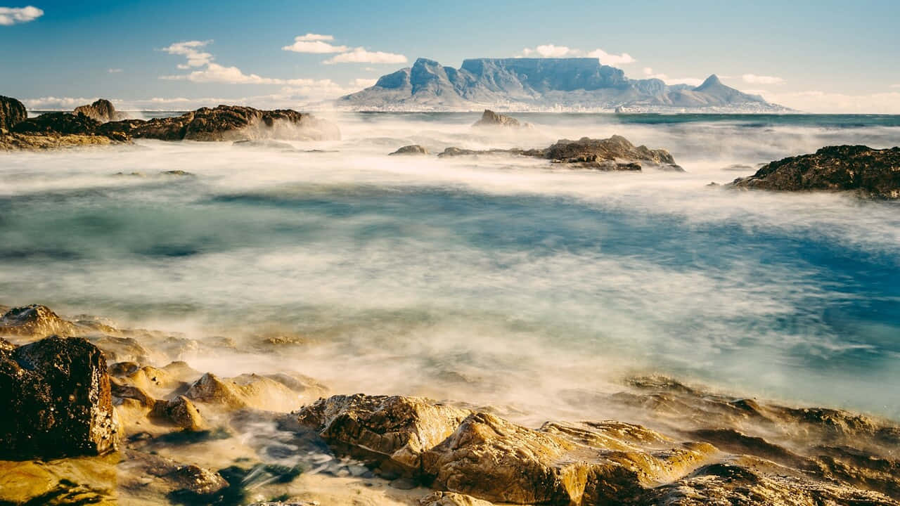 A Rocky Shore With Waves And Mountains In The Background