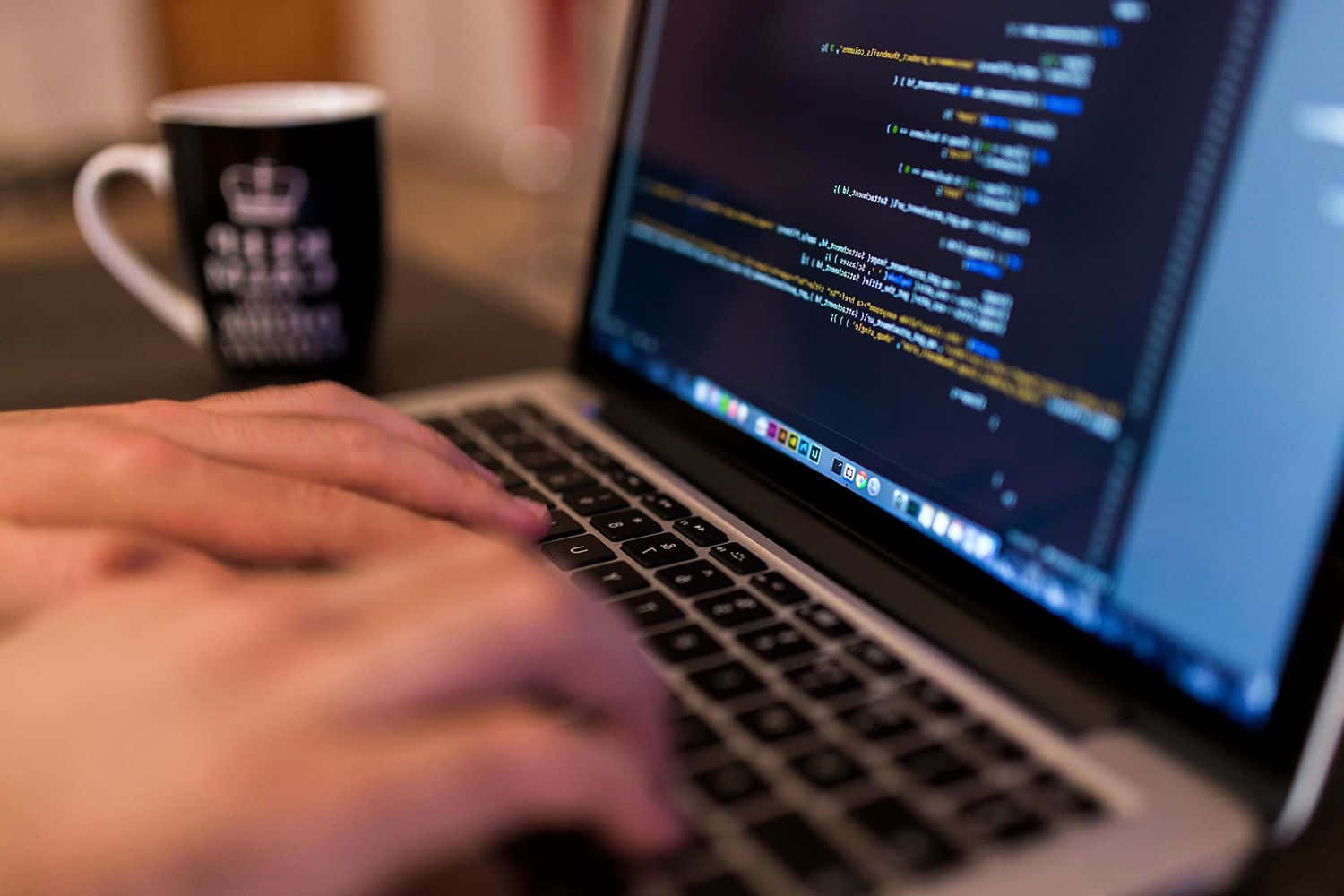 A programmer hard at work Picture — Hands typing on a laptop keyboard with a black mug nearby and code visible on the screen.