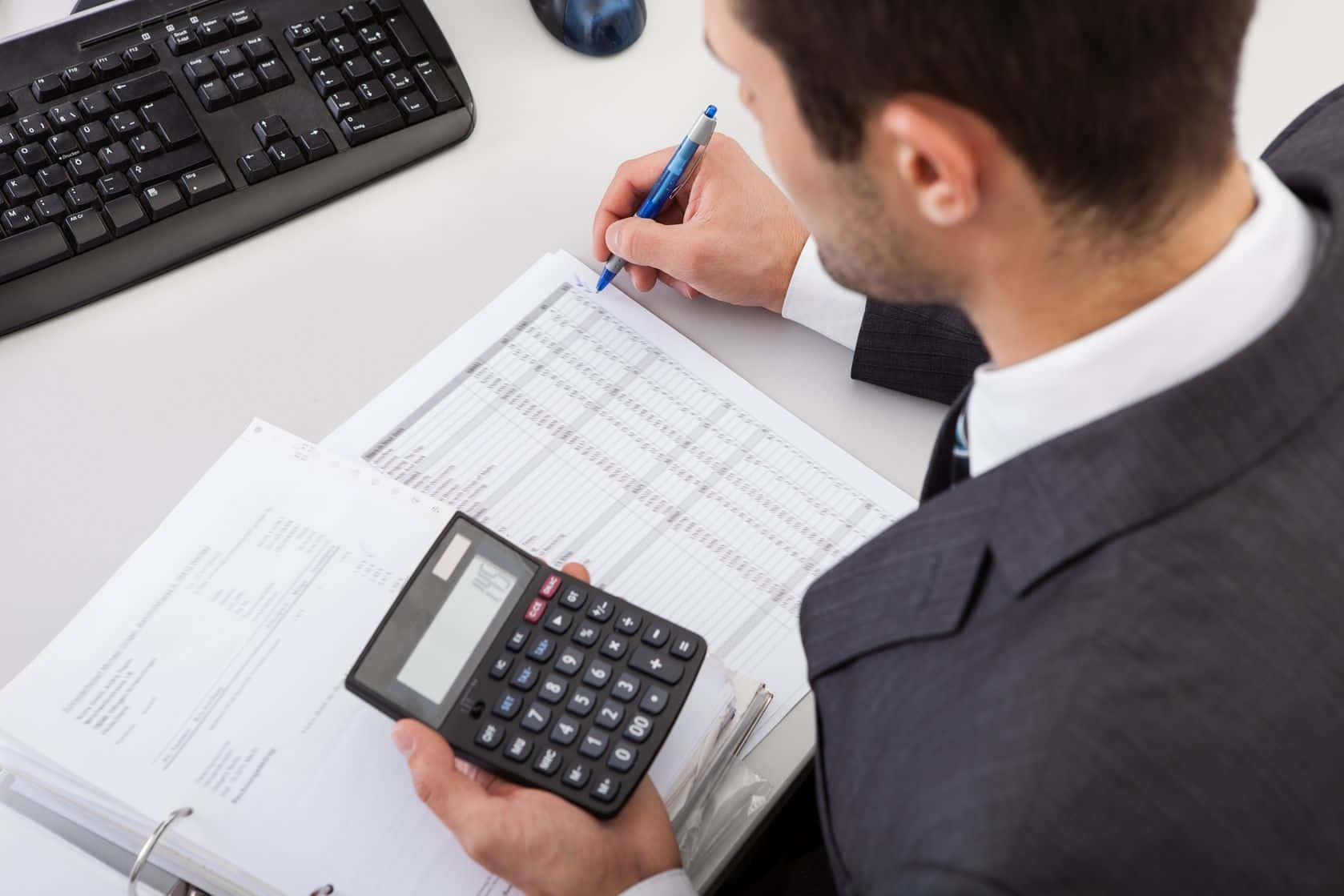 A Man In A Suit Is Using A Calculator At His Desk