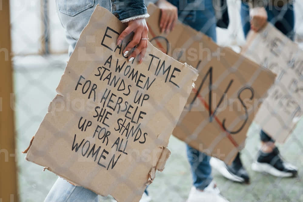 Women Holding Signs That Say Every Time A Woman Stands Up For Herself, She Stands Up For Women Wallpaper