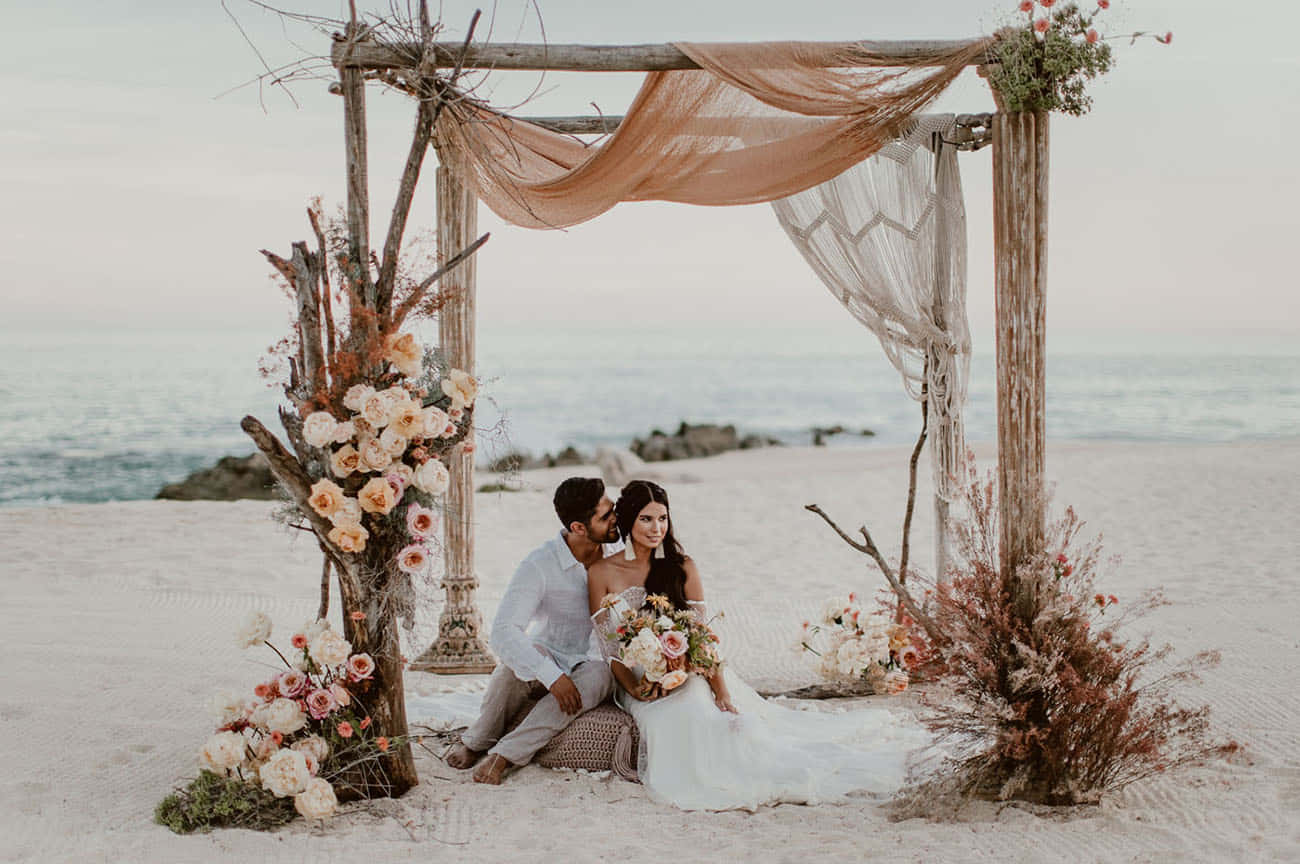 A Bride And Groom Sitting On A Wooden Arch On The Beach
