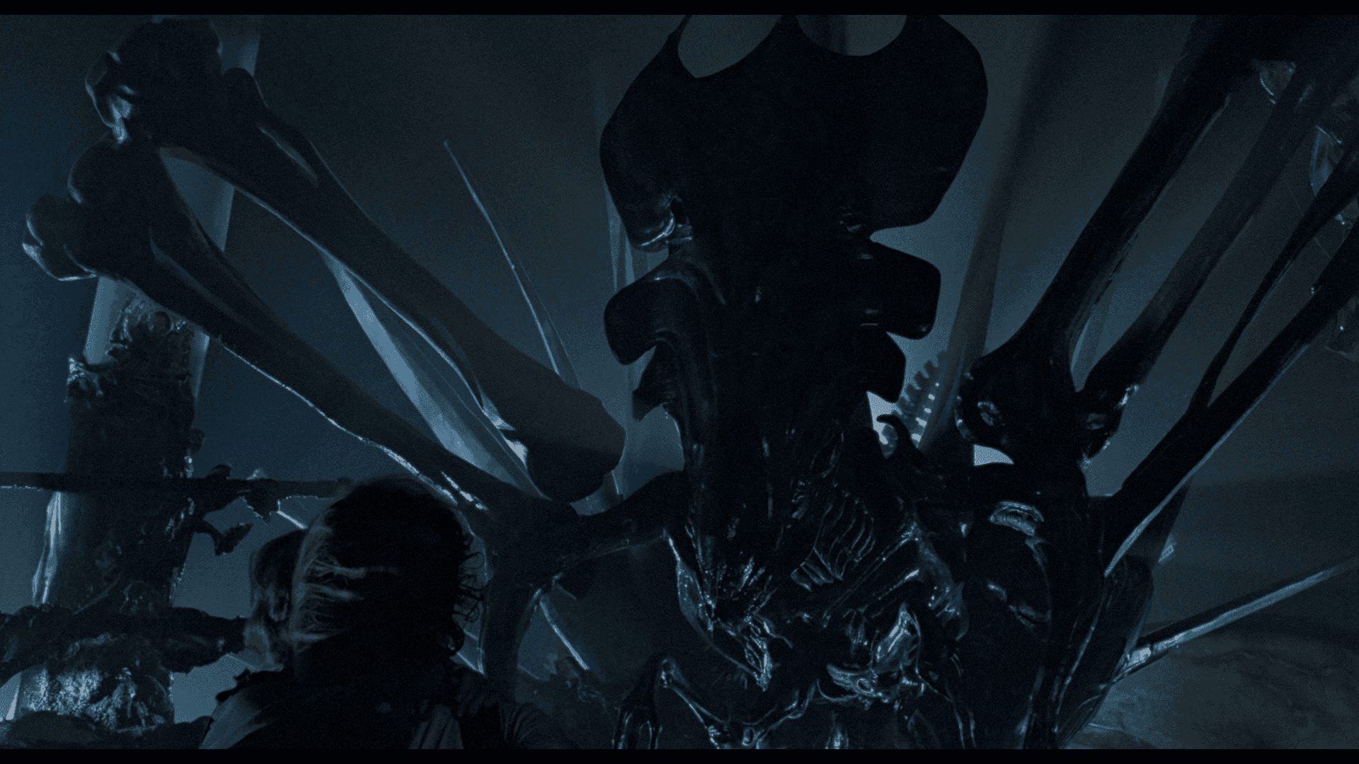 aliens - the movie - screenshot Background — A towering alien creature with biomechanical limbs looms over a terrified human in a dimly lit, industrial setting.