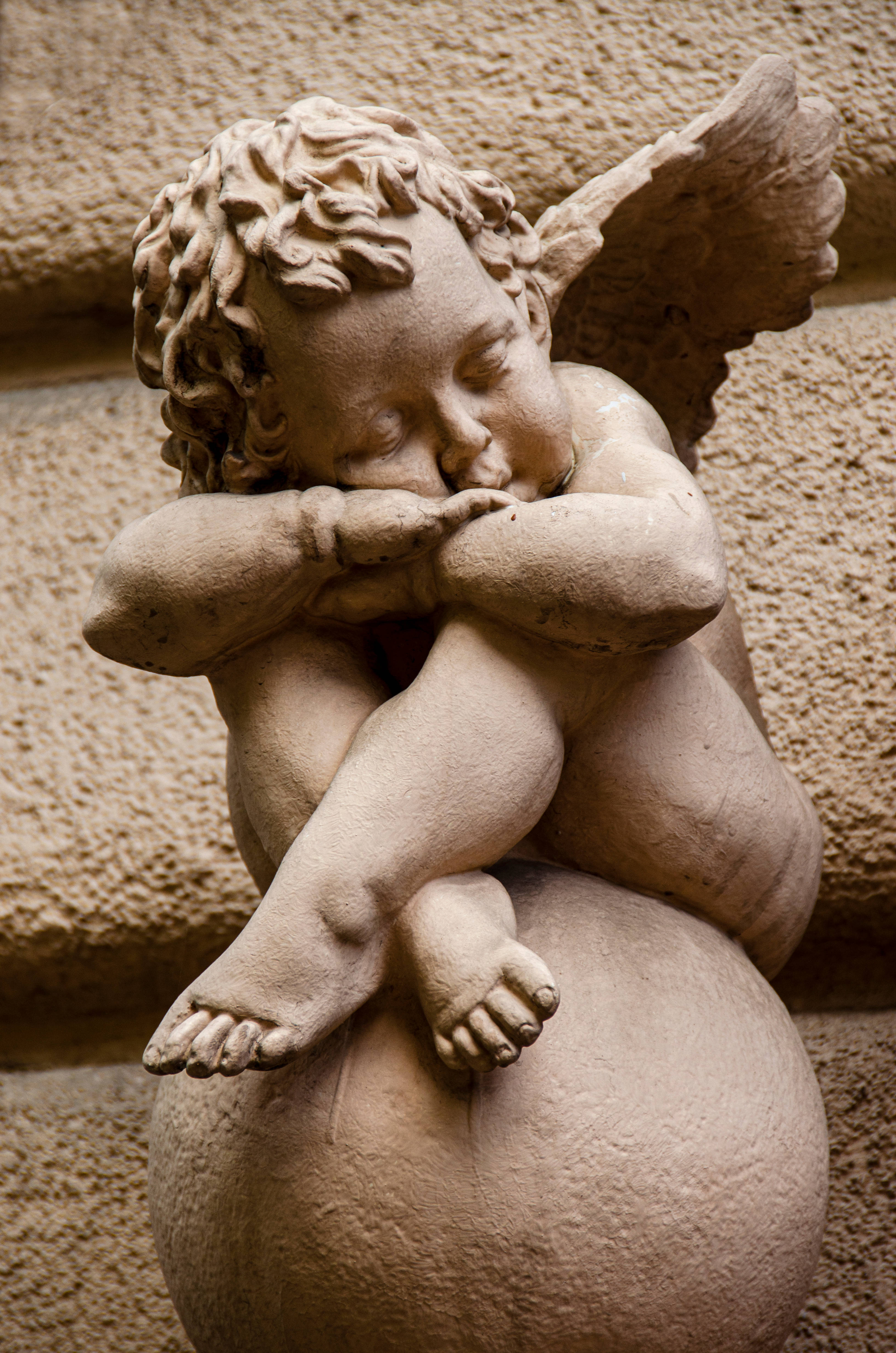Angel Wallpaper — A stone cherub statue with curly hair and wings sleeps curled on a sphere against a textured wall.