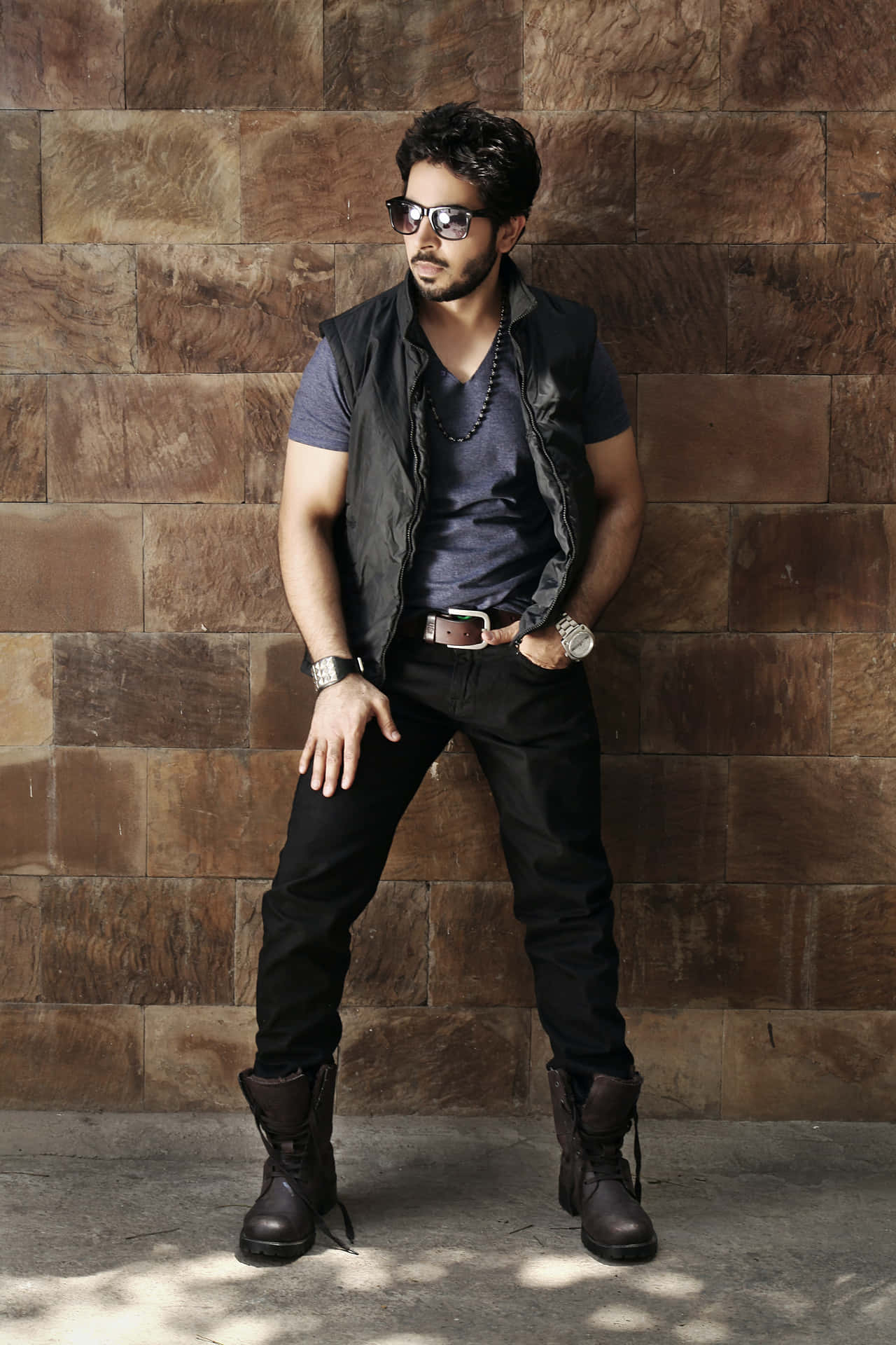 A Man In A Black Vest And Jeans Leaning Against A Brick Wall