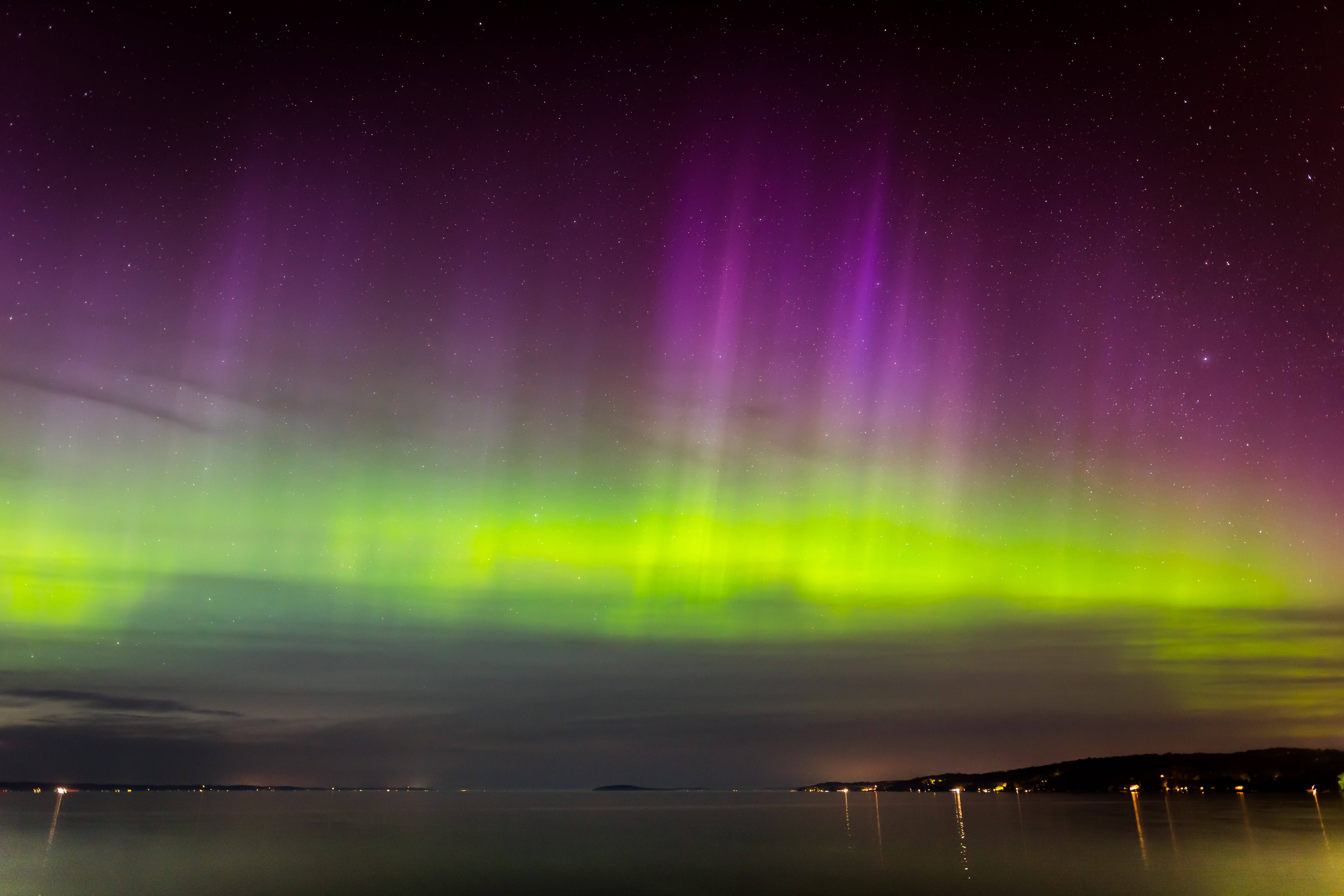 Green And Violet Northern Lights Wallpaper — Vibrant green and purple auroras dance across a starry night sky above a calm reflective water surface with distant lights.