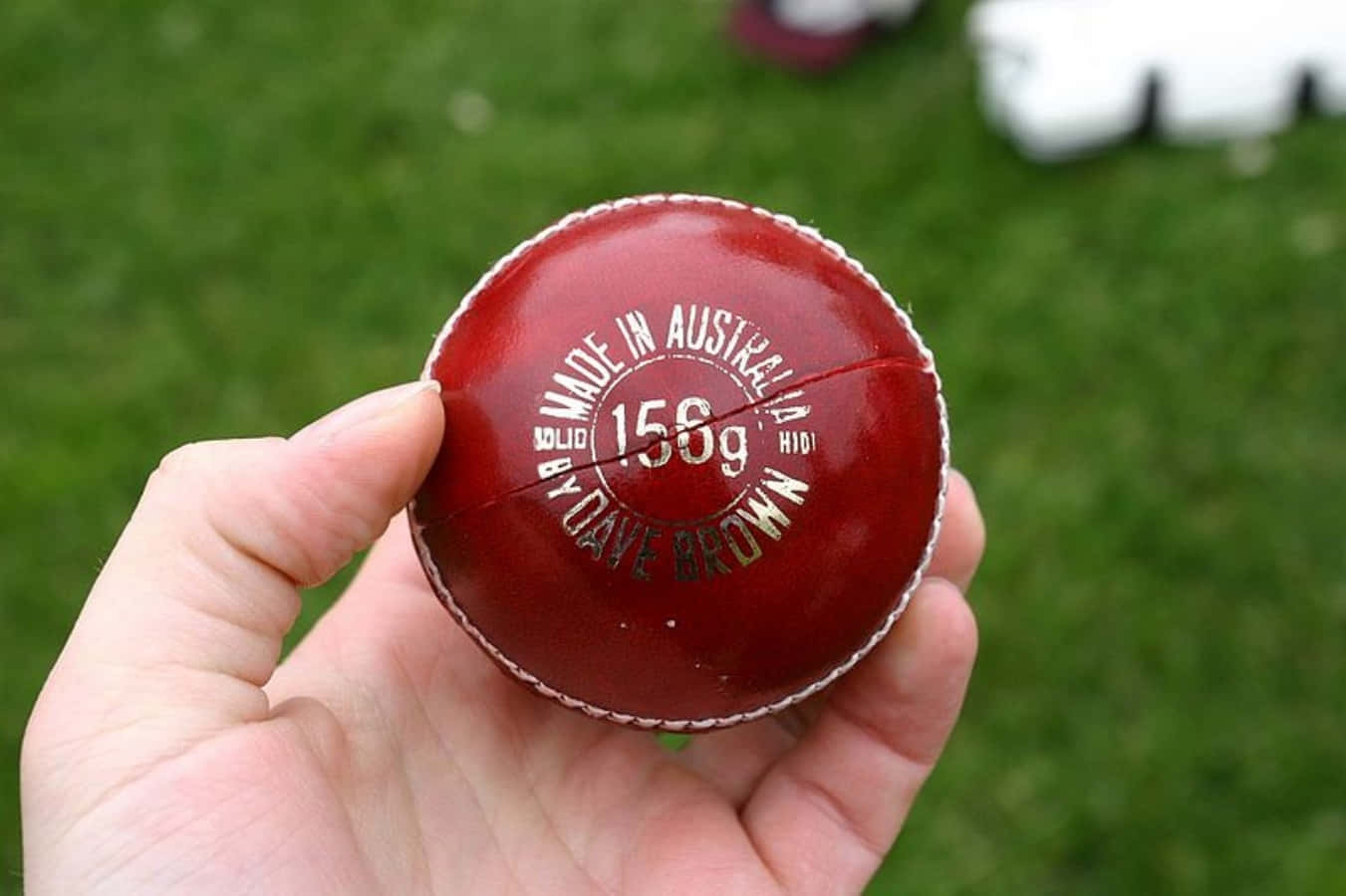 A Person Holding A Red Cricket Ball In Front Of A Field