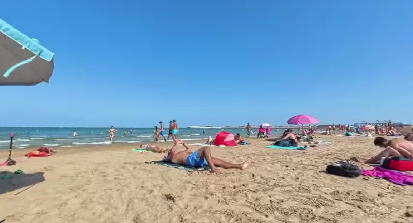 beach PNG — People relax on a sunny beach under clear blue skies with colorful umbrellas and gentle ocean waves.
