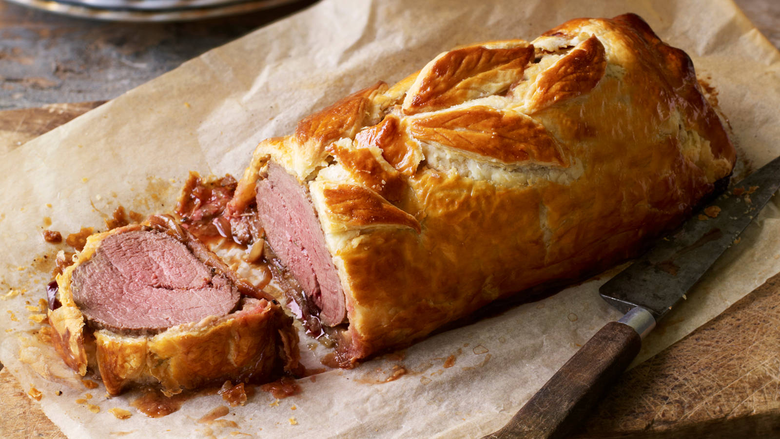 Beef Wellington Juicy Knife Wallpaper — A golden-brown beef Wellington with flaky pastry and rich sauce rests on parchment paper beside a carving knife.
