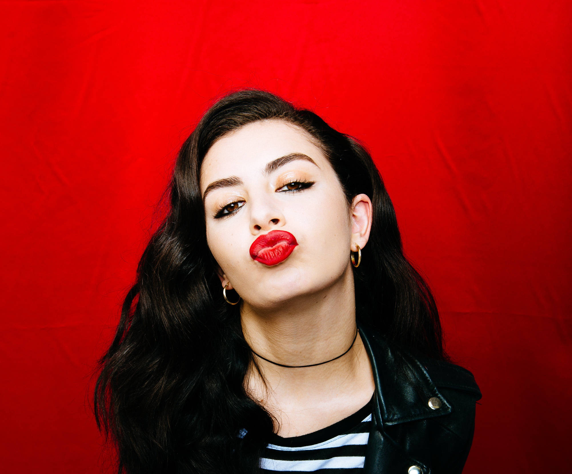 Kiss From Charli Xcx Wallpaper — A woman with dark hair and bold red lips blows a kiss against a vivid red backdrop wearing a black leather jacket.