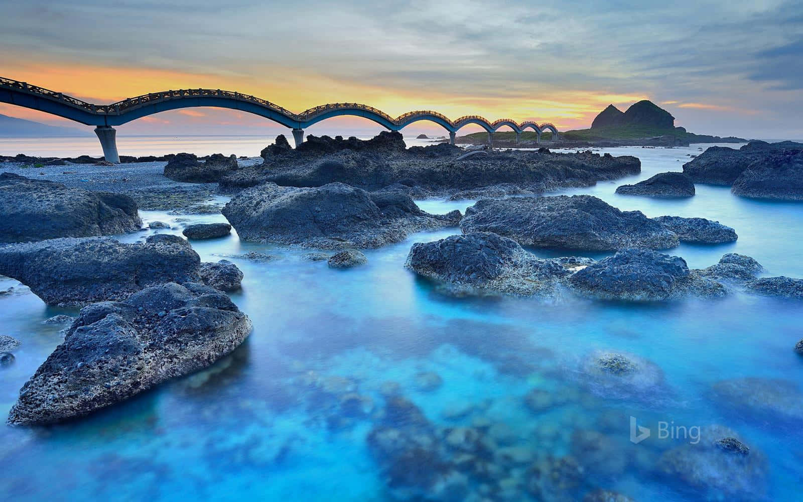 A Bridge Over The Ocean With Rocks In The Background