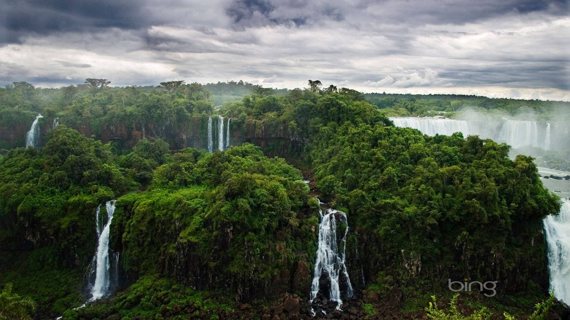 Take in a peaceful view of a green forest, surrounded by majestic waterfalls Wallpaper