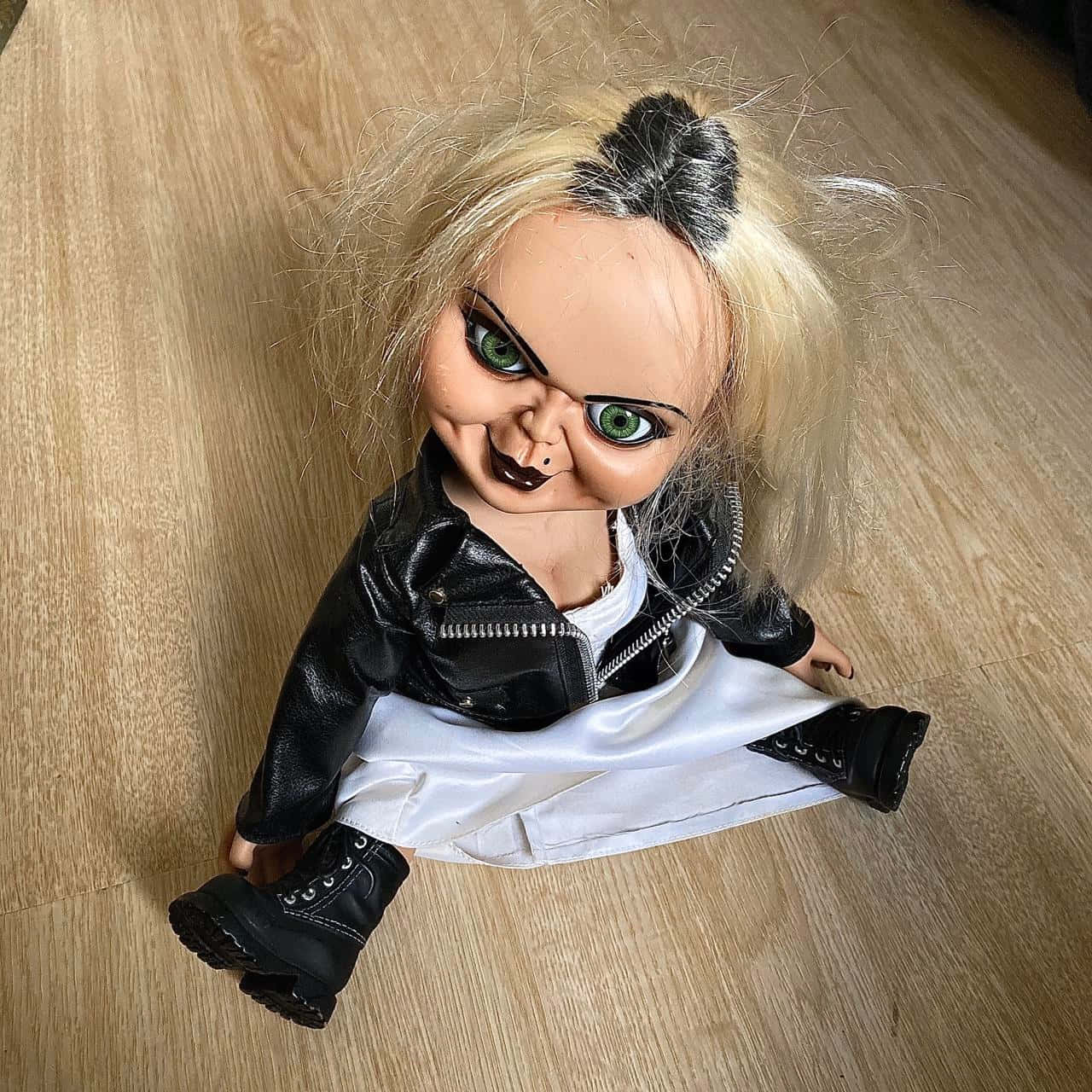 Brideof Chucky Doll Image Wallpaper — A creepy doll with green eyes and black leather jacket sits on a wooden floor.