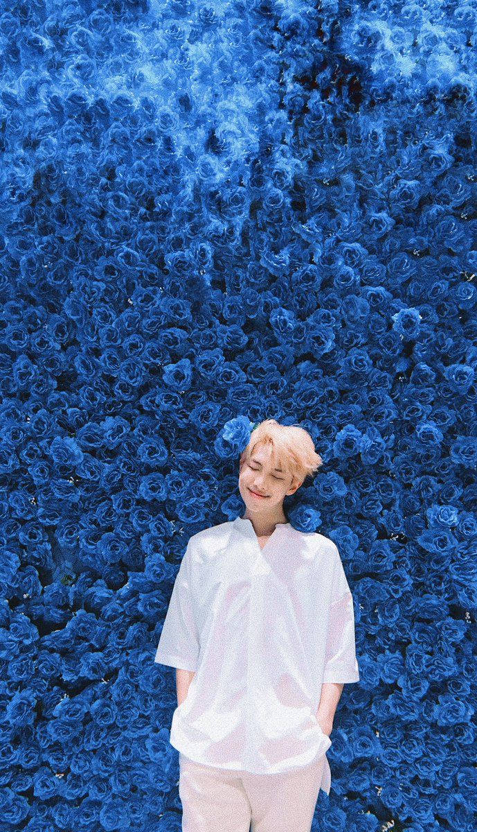 BTS member standing in front of a wall full of blue flowers. 