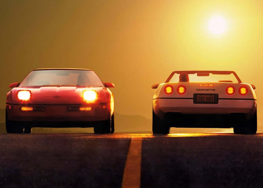 Two Sports Cars Are Driving Down The Road At Sunset