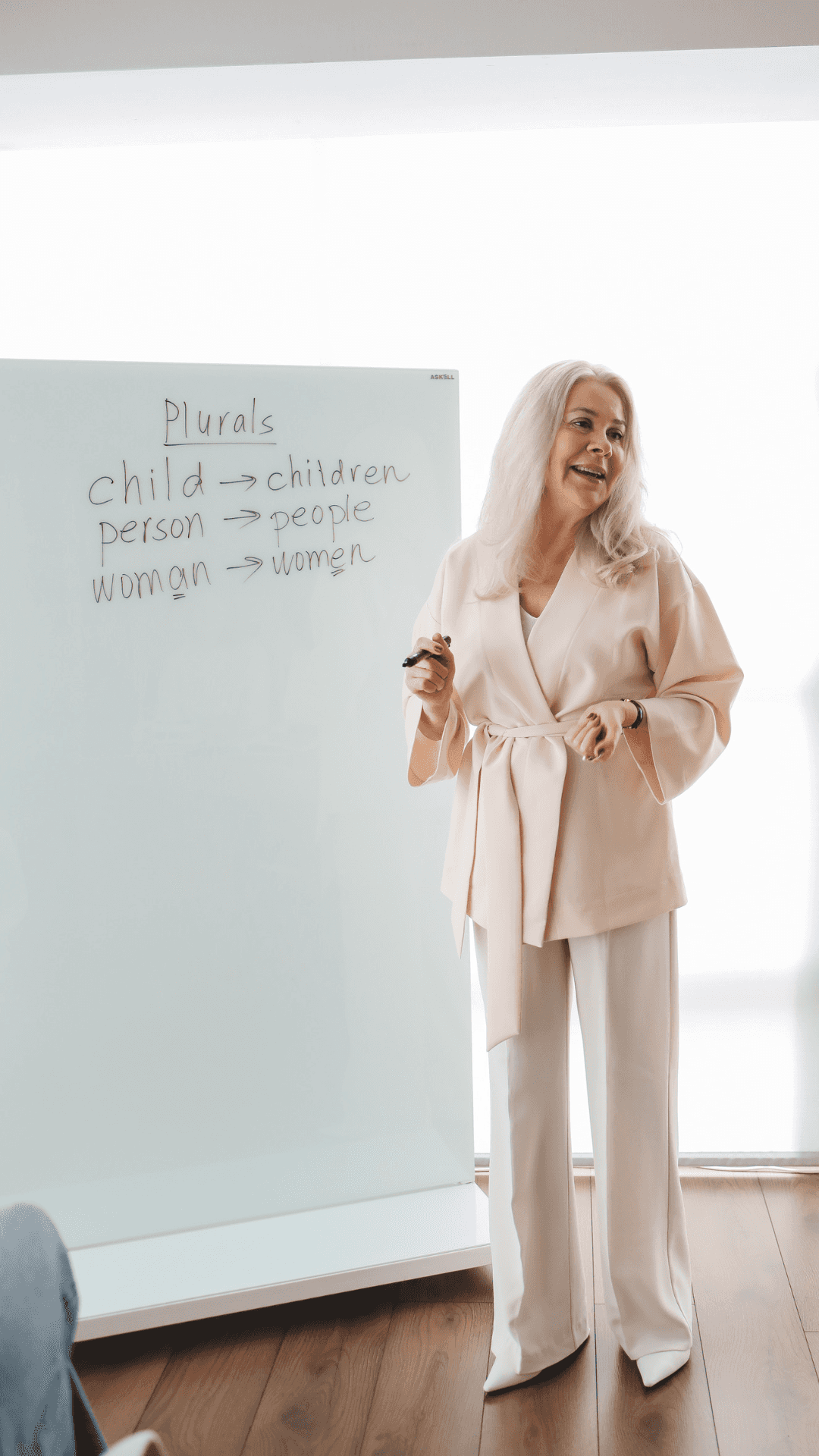 Caption: Interactive Teaching Session in Progress Background — A smiling woman in beige attire teaches grammar on a whiteboard in a bright modern classroom.